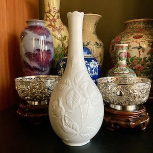 May include: A white porcelain vase with a floral relief design, featuring a slender neck and a flared rim. The vase is surrounded by other decorative vases and silver bowls on wooden stands, creating a collection of antique home decor.