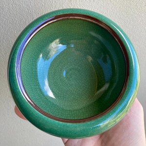 May include: A round, green ceramic bowl with a crackle glaze. The bowl has a dark brown rim and a glossy finish. The interior is a lighter shade of green. The bowl is held in a hand.