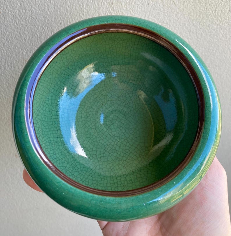 May include: A round, green ceramic bowl with a crackle glaze. The bowl has a dark brown rim and a glossy finish. The interior is a lighter shade of green. The bowl is held in a hand.