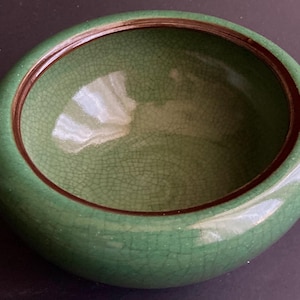 May include: A round, green ceramic bowl with a crackle glaze. The bowl has a dark brown rim and a smooth, glossy exterior. The interior of the bowl is a lighter green color.