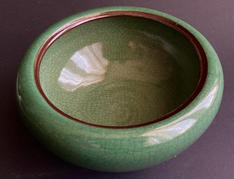 May include: A round, green ceramic bowl with a crackle glaze. The bowl has a dark brown rim and a smooth, glossy exterior. The interior of the bowl is a lighter green color.