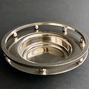 May include: A silver-colored, circular serving dish with a raised, rounded rim and a handle-like structure. The dish has a reflective surface and small, spherical accents around the rim. The design is simple and elegant.