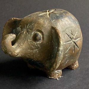 May include: A small, brown, hand-sculpted elephant figurine with a textured, weathered surface. The elephant features carved star-like patterns on its head and side, large ears, a curved trunk, and four short, sturdy legs, all in a rustic finish.