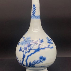 May include: A white ceramic vase with a bulbous body and a long, slender neck. The surface is decorated with hand-painted blue floral and botanical motifs, including leafy branches and blossoms, featuring a flared base and a narrow rim opening.