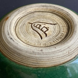 May include: Close-up of the bottom of a green ceramic bowl. The base is a light beige color with concentric circles and a carved symbol. The bowl's exterior is a glossy, dark green.