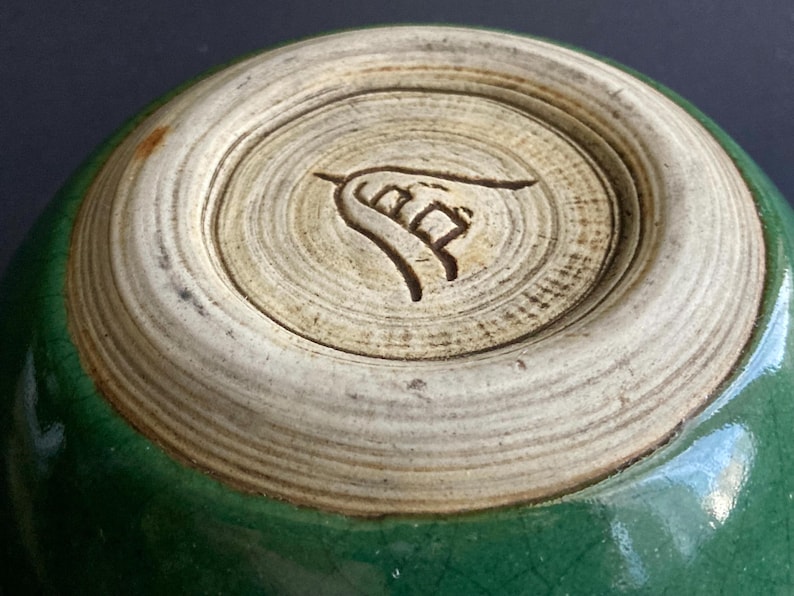 May include: Close-up of the bottom of a green ceramic bowl. The base is a light beige color with concentric circles and a carved symbol. The bowl's exterior is a glossy, dark green.