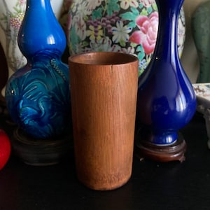 Puede incluir: Una taza cilíndrica de madera de color marrón claro natural. La taza está hecha de listones de madera verticales y tiene una superficie lisa y sin adornos. La taza se exhibe frente a una colección de jarrones decorativos.