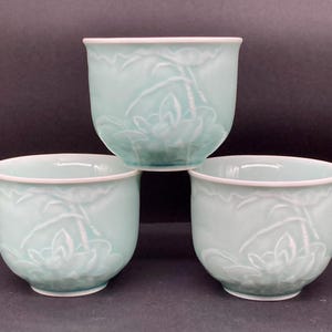 May include: Three pale mint green ceramic tea cups arranged in a pyramid formation. Each cup features a raised, embossed floral design of lotus flowers and stems on the exterior surface, with a smooth, rounded rim and a solid base.