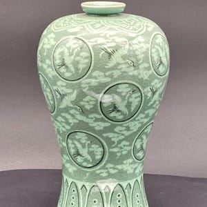 May include: A light green celadon vase with a crackle glaze finish. The body features circular medallions containing crane illustrations, surrounded by cloud motifs. The neck has a floral pattern, and the base is decorated with vertical petals.