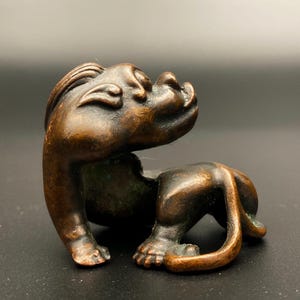 May include: A small, dark bronze-toned metal figurine of a stylised creature with a smooth, curved body, a prominent head with defined facial features, and a long tail that curls around its hind legs, featuring a weathered, aged patina finish.