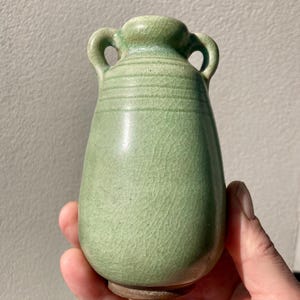 May include: A small, light green ceramic vase with a bulbous body and two arched handles near the narrow neck. The surface features a distinct crackle glaze finish and horizontal incised lines around the upper section, with a brown base.
