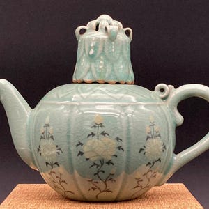 Op de afbeelding: Een lichtgroene keramische theepot met een craquelé glazuurafwerking, voorzien van een geribbelde behuizing en een bijpassend deksel met een decoratieve top. De buitenkant is versierd met geschilderde bloemmotieven in wit en donkergrijs.