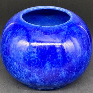 May include: A spherical ceramic bowl with a glossy finish, featuring a vibrant cobalt blue glaze with subtle variations in tone and texture across its rounded surface. The bowl has a wide, circular opening at the top and a smooth, curved body.