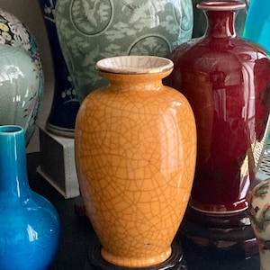 Könnte beinhalten: Eine orangefarbene Keramikvase mit Craquelé-Glasur, auf einem dunklen Holzständer. Die Vase hat einen goldenen Rand und ist von anderen dekorativen Vasen in Blau-, Grün- und Rottönen umgeben.