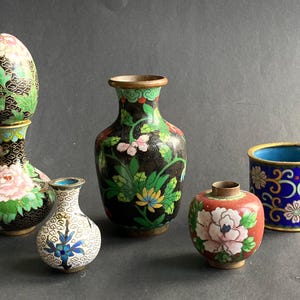 May include: A collection of five cloisonné items including a black vase topped with a floral egg, a smaller white vase with blue flowers, a medium black vase with yellow and pink flowers, a small red jar with a pink flower, and a blue cylindrical pot.