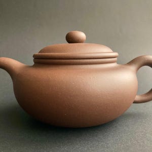 Könnte beinhalten: Eine braune Keramik-Teekanne mit rundem Körper, gebogenem Ausguss und Schlaufengriff. Der Deckel hat einen kleinen, runden Knopf. Die Teekanne hat ein mattes Finish und steht auf einer dunklen Oberfläche. Tee- und Küchenartikel.