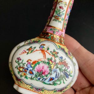 May include: A small, white porcelain vase with a bulbous body and long neck. The vase is hand-painted with colorful birds, flowers, and foliage. Gold trim accents the edges. The vase is held in a hand.