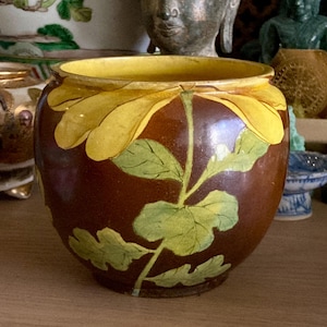 May include: A round ceramic bowl with a glossy brown glaze, featuring a large yellow flower painted on the front with green leaves and a stem extending downwards. The interior of the bowl is a solid yellow colour, and the rim is smooth and rounded.