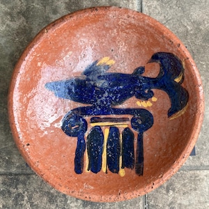 May include: A terracotta plate with a blue and yellow painted design of a fish on a column. The fish is facing left and has a yellow fin. The column is painted with yellow and blue stripes.