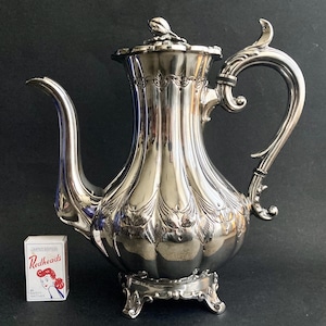 May include: A silver-colored, ornate coffee pot with a curved spout and handle. The pot has a decorative lid and sits on a small, three-legged base. A box of Redheads safety matches is next to the pot.