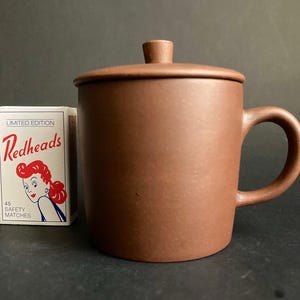 以下が含まれることがあります： 茶色のセラミックマグカップ、お揃いの蓋とハンドル付き。マグカップの隣には、ヴィンテージイラストの赤毛の女性が描かれた限定版の「Redheads」マッチ箱があります。マグカップの高さは約10cmです。