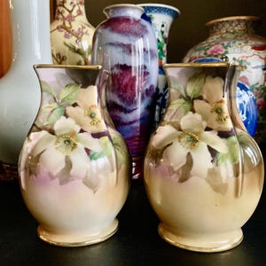 Puede incluir: Un par de jarrones de porcelana vintage con un diseño floral. Los jarrones son de color crema con un borde dorado y presentan flores blancas con hojas verdes.