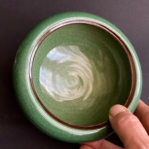 May include: A round, green ceramic bowl with a crackle glaze interior and a dark brown rim. The bowl is held by a hand, showcasing its size and shape. The bowl's exterior is a solid, glossy green.