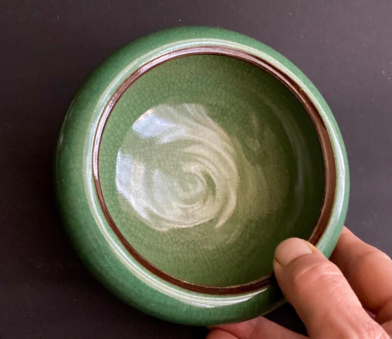 May include: A round, green ceramic bowl with a crackle glaze interior and a dark brown rim. The bowl is held by a hand, showcasing its size and shape. The bowl's exterior is a solid, glossy green.