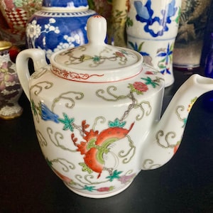 Pode incluir: Uma chaleira de porcelana branca com um design floral em tons de vermelho, verde, azul e laranja. A chaleira tem um bico curvo e uma alça no lado.