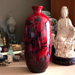 Puede incluir: Un jarrón alto de cerámica roja con un acabado brillante. El jarrón presenta una escena oscura y detallada de un bosque con ciervos. El jarrón es un artículo decorativo, probablemente para la decoración del hogar, y se encuentra sobre una superficie de madera.