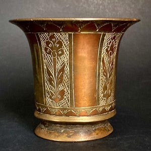May include: A copper-coloured metal vase with etched floral and geometric patterns. The vase has a flared rim and a pedestal base. The etched designs are in a lighter gold colour, creating a contrast against the copper background. Likely an antique or decorative piece.