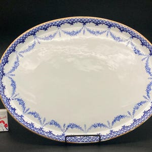 May include: An oval white ceramic serving platter featuring a decorative blue floral garland pattern along the rim and a thin gold-colored edge. A small rectangular matchbox with the text Redheads is positioned to the left for scale.