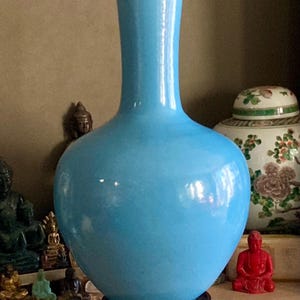 May include: A bright sky blue ceramic vase with a bulbous body and a slender, elongated neck. The vase features a smooth, glossy finish and is displayed resting on a dark, circular wooden stand, providing a stable base for the vessel.