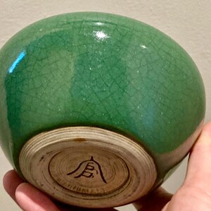 May include: A green ceramic bowl with a crackle glaze. The bowl has a rounded shape and a light-colored, unglazed base with a black symbol. The bowl is held in a hand.