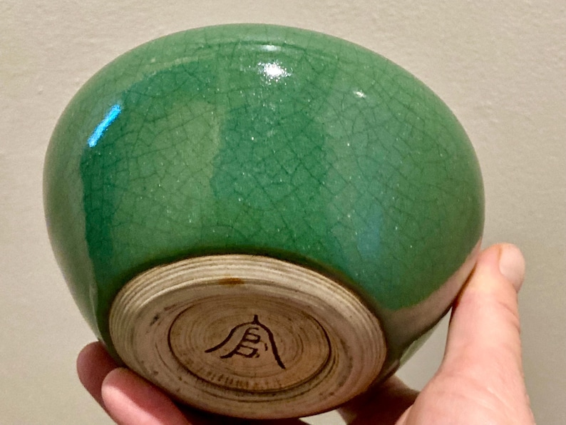 May include: A green ceramic bowl with a crackle glaze. The bowl has a rounded shape and a light-colored, unglazed base with a black symbol. The bowl is held in a hand.
