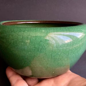 May include: A green ceramic bowl with a crackle glaze. The bowl has a rounded shape with a dark brown interior rim. The exterior is a vibrant green color. The bowl is held in a hand.