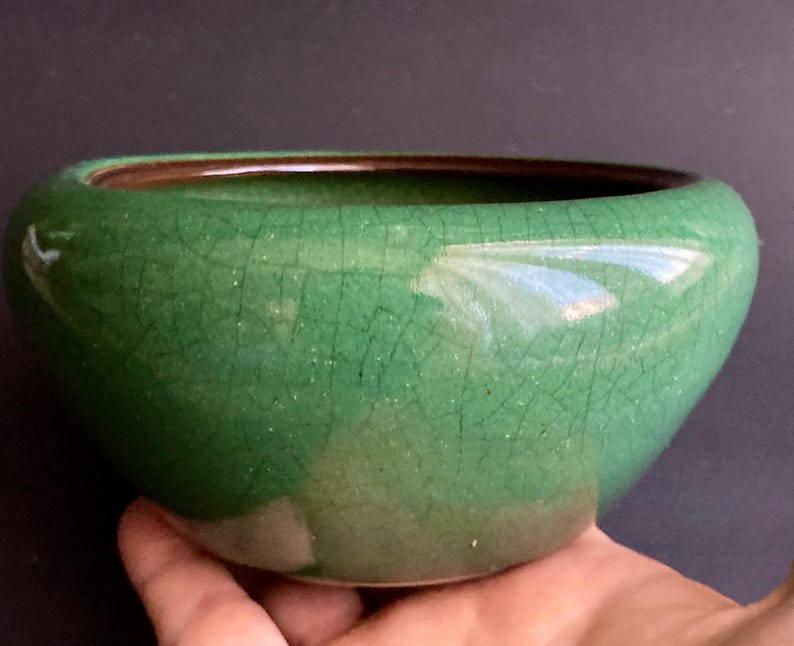 May include: A green ceramic bowl with a crackle glaze. The bowl has a rounded shape with a dark brown interior rim. The exterior is a vibrant green color. The bowl is held in a hand.