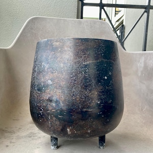 Peut inclure: Un pot en métal sombre et vieilli, avec un corps arrondi et trois courtes pattes. Le pot a une finition vieillie avec des nuances de noir, de brun et des touches de bleu. Le dessus ouvert suggère qu'il est conçu pour les plantes.