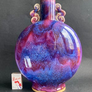 May include: A round, glazed ceramic vase with a vibrant blend of purple, blue, and red hues. The vase features two decorative handles and a flared base. A box of "Redheads" matches is next to the vase.