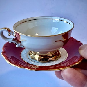 May include: A white teacup and saucer set with gold trim. The saucer is a deep red color with a white floral design. The teacup has a white interior and a gold rim.