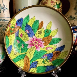 May include: A decorative ceramic plate with a floral design. The plate features a central pink flower surrounded by green, yellow, blue, and brown leaves. The rim is trimmed with gold. The plate is displayed on a clear stand.