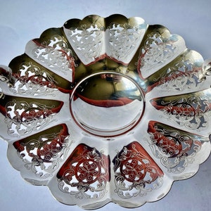 May include: A silver serving dish with intricate cut-out designs and two handles. The dish has a scalloped edge and a shiny, reflective surface.