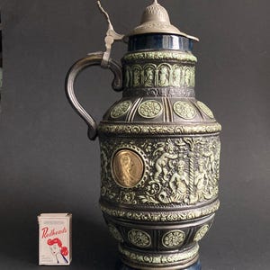 Op de afbeelding: Een antieke keramische bierpul met een metalen scharnierend deksel en handvat. De pul is donkergroen en zwart met lichtgroene reliëfontwerpen, waaronder figuren en medaillons. Een doosje Redheads lucifers staat ernaast.