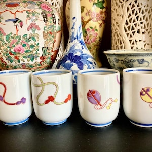 May include: Four white teacups with blue rims and colourful floral and geometric designs. The cups are arranged in a row on a black surface.