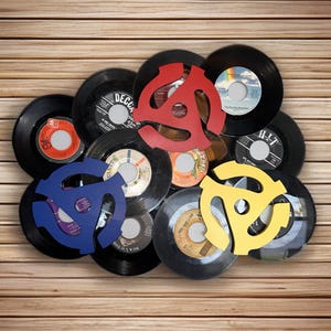 May include: A collection of black vinyl records with colorful 45 RPM record adapters in red, blue, and yellow. The records are arranged on a wooden background, showcasing a vintage music theme.