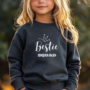 Puede incluir: Una sudadera gris oscuro con un gr&aacute;fico blanco que dice "Bestie Squad" con una corona sobre el texto.