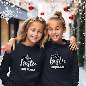 Puede incluir: Dos chicas con sudaderas negras con una corona blanca y el texto "Bestie Squad" en ellas. Las chicas est&aacute;n de pie frente a un fondo borroso de una calle de la ciudad con decoraciones navide&ntilde;as.