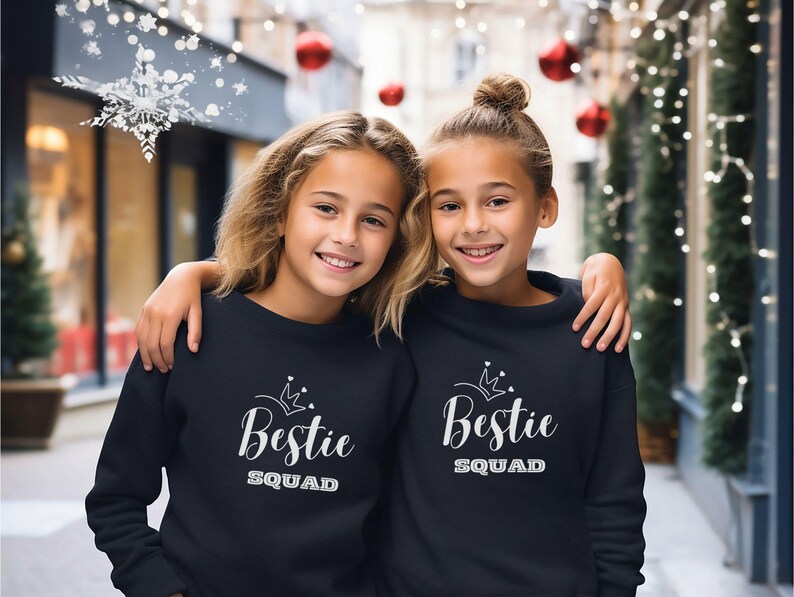 Puede incluir: Dos chicas con sudaderas negras con una corona blanca y el texto "Bestie Squad" en ellas. Las chicas est&aacute;n de pie frente a un fondo borroso de una calle de la ciudad con decoraciones navide&ntilde;as.