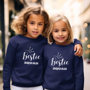 Puede incluir: Dos chicas con sudaderas azules marino a juego con una corona blanca y el texto "Bestie Squad".