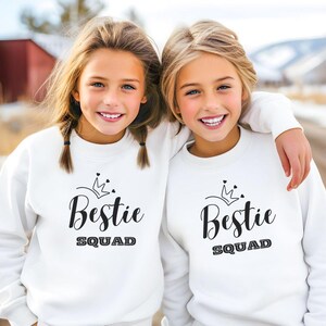Puede incluir: Dos chicas con sudaderas blancas con el texto "Bestie Squad" y un gr&aacute;fico de corona. Las chicas sonr&iacute;en y miran a la c&aacute;mara.